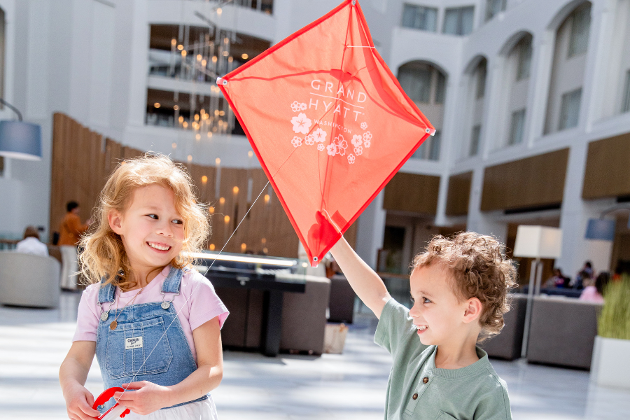 Two smiling children play with a red Grand Hyatt Washington kite in the hotel's bright and modern atrium.