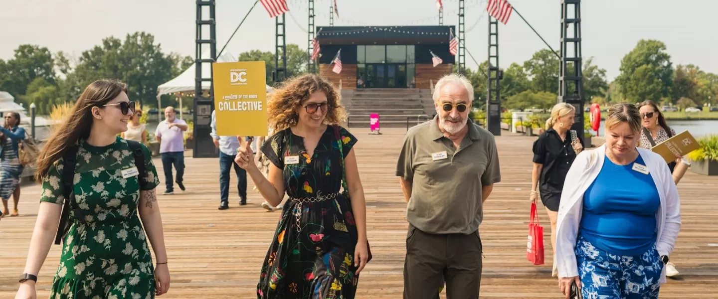 A group strolls along a sunny boardwalk, led by a smiling participant holding a "Collective" sign, under festive string lights and American flags.