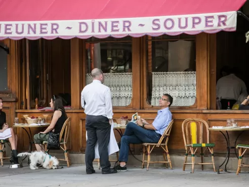 Diners relax with drinks and meals at an outdoor French café with lace-curtained windows and pink awning on a DC street.