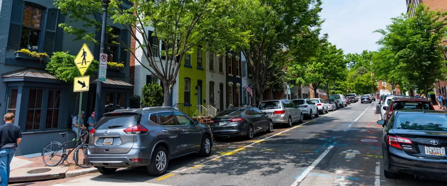 A tree-lined residential street in Georgetown with parked cars, colorful rowhouses, and pedestrians walking along the sidewalk on a sunny day.