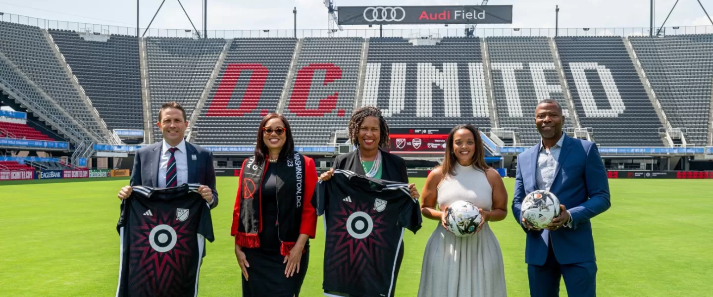 A group of five people stands on the field at Audi Field stadium holding soccer jerseys and balls, with "D.C. UNITED" visible in bold red and white lettering across the empty stands behind them.