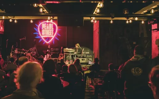 An intimate live music performance at Pearl Street Warehouse, with a neon sign in the shape of a guitar pick glowing on the back wall. The audience is seated, watching the performer on stage, with warm lighting overhead.