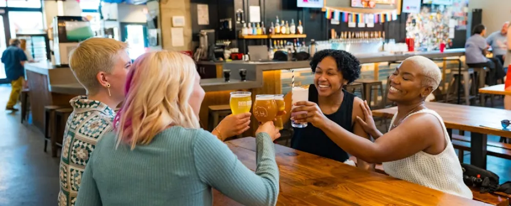 Two couples smile and clink their beers together at a brewery. 