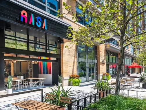 The exterior of Rasa restaurant in Mount Vernon Square features bright modern decor and a sunny outdoor patio with seating and greenery.